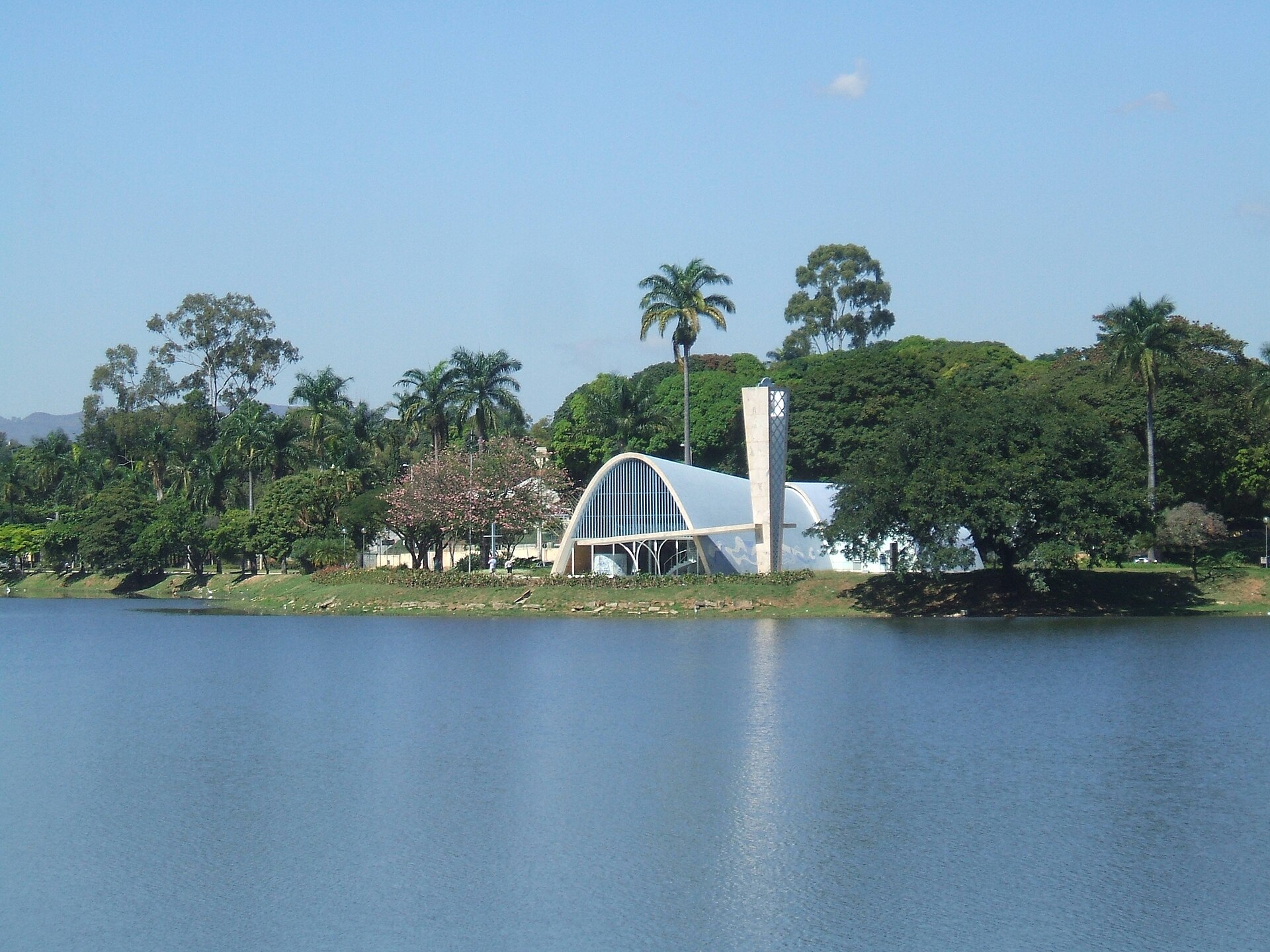 Igreja São Francisco de Assis, com as abóbadas brancas e a torre, vista do outro lado da Lagoa da Pampulha, entre palmeiras e árvores