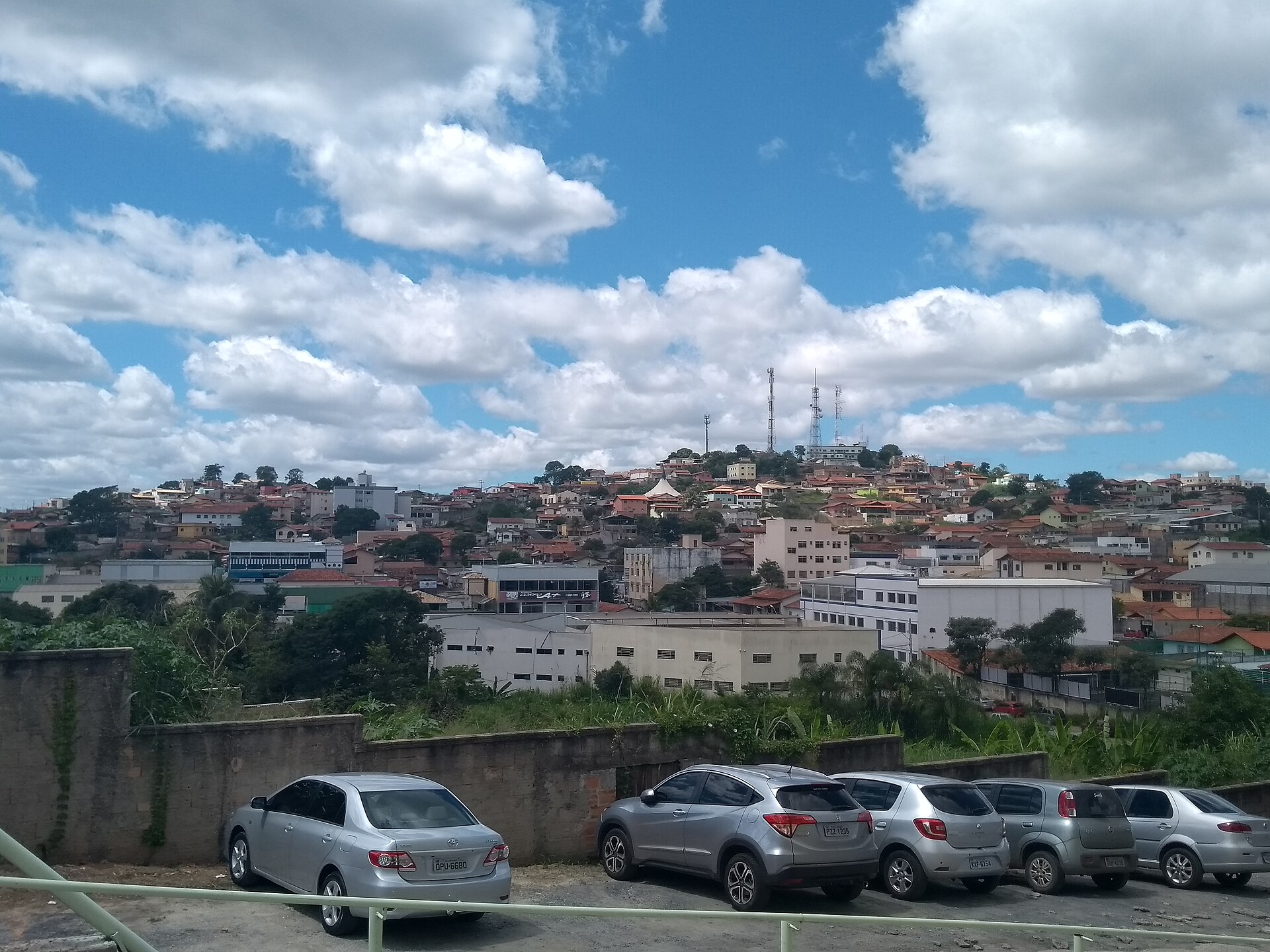 Vista de Ribeirão das Neves: casas e prédios espalhados pelo morro, com torres de antena no alto e céu azul com nuvens